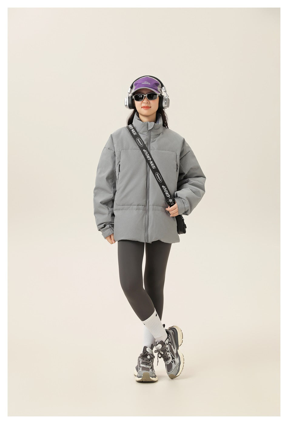 Model wearing gray puffer jacket zipped up with crossbody strap, headphones, sunglasses, leggings and sneakers
