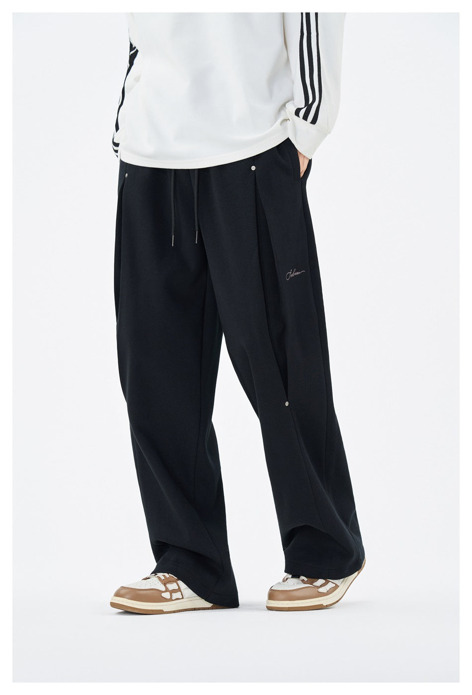 Three-quarter front view of a model wearing loose black wide-leg trousers with drawstrings and snap details.