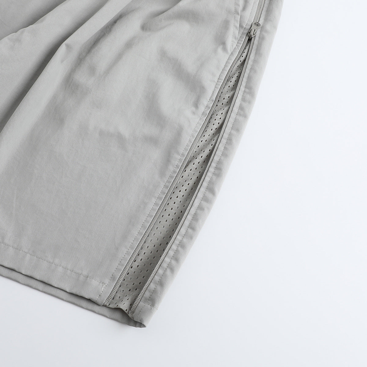 Close-up of a side zipper opened to reveal a perforated inner panel on light gray shorts.