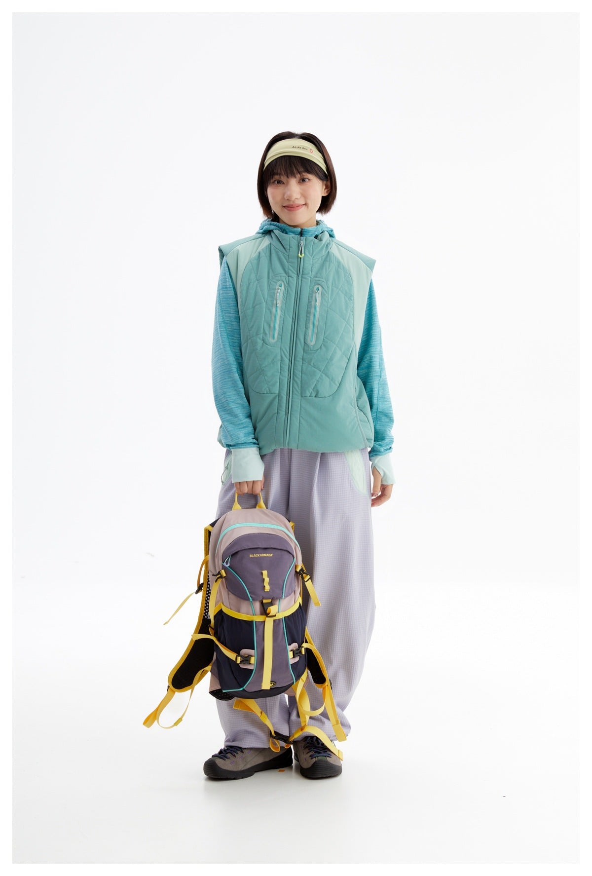 Front view of a model wearing a teal quilted zip vest with two chest pockets and a hood, paired with lavender pants.