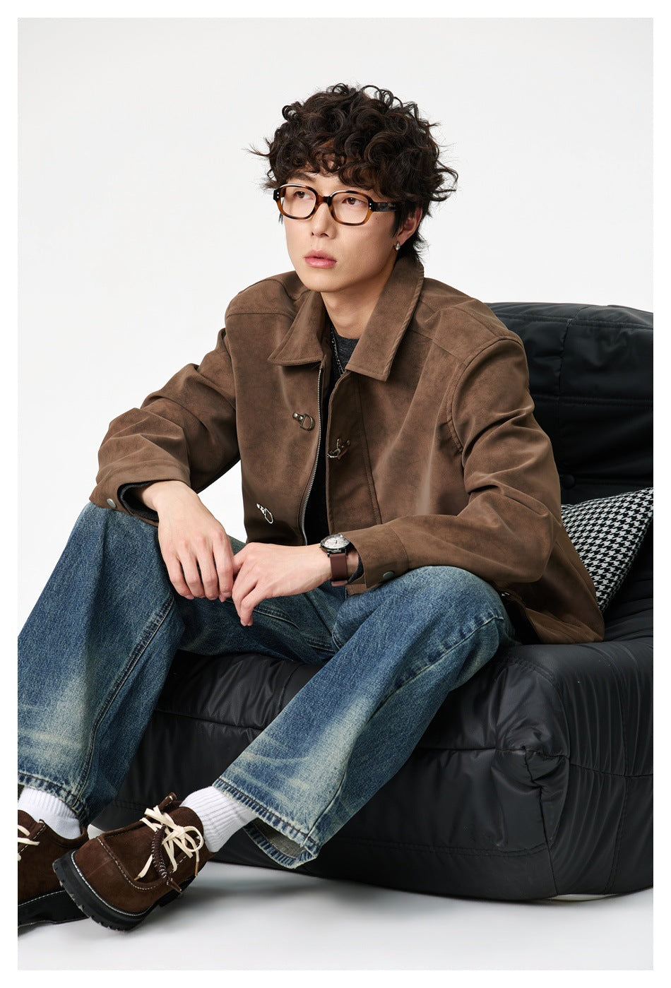 Person seated on a black chair wearing a brown suede-look jacket with metal hook closures, blue jeans and brown shoes