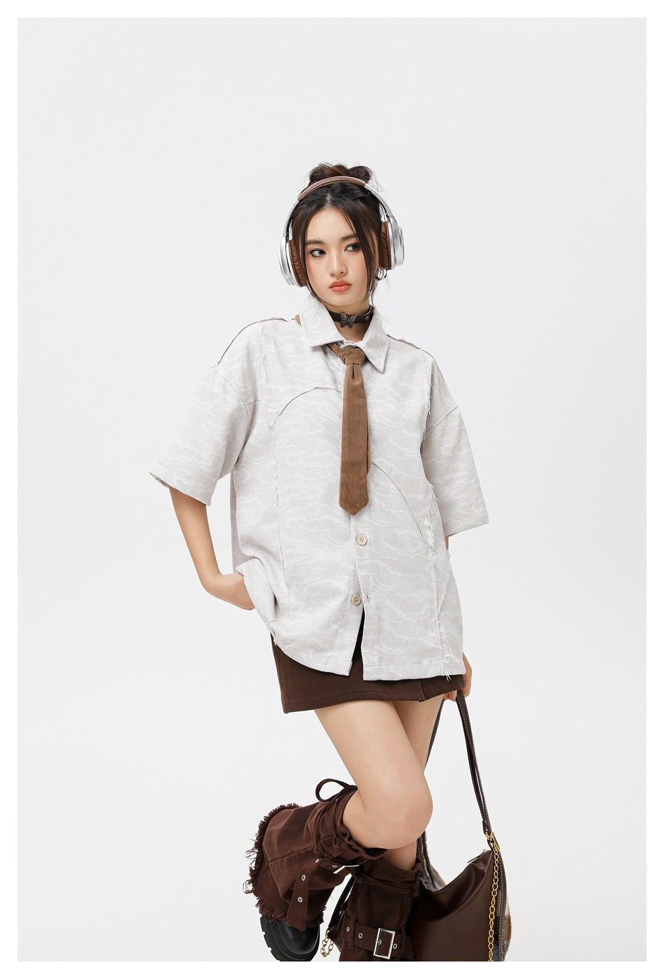 Model poses in an oversized pale gray patterned short-sleeve button-up shirt, brown tie, and dark brown skirt.