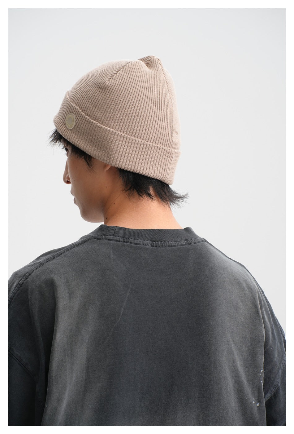 Rear view of a person wearing a ribbed beige cuffed beanie with a small round patch and a charcoal T-shirt.