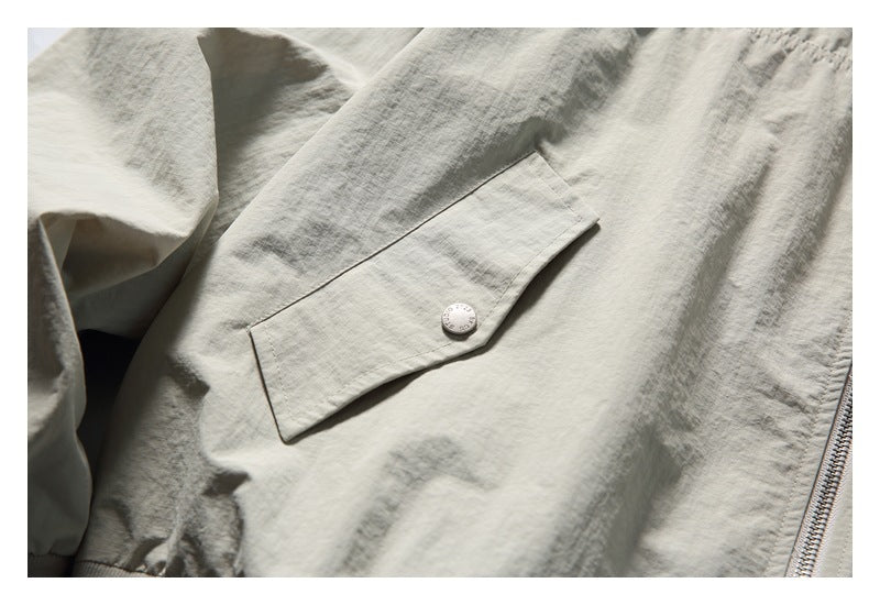 Close-up of a buttoned flap pocket on a light stone-gray jacket, with part of the front zipper visible.