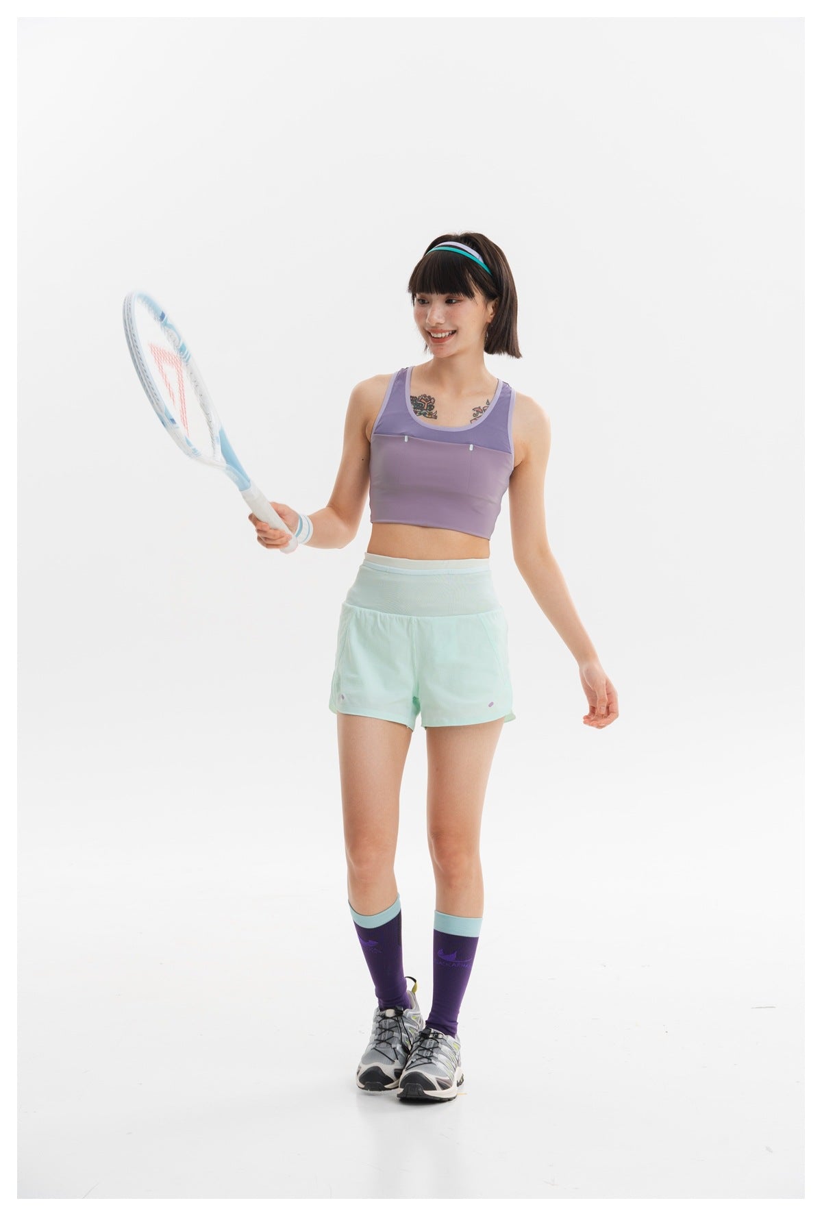 Woman in a lavender sports bra and mint shorts holding a tennis racket against a white studio background.