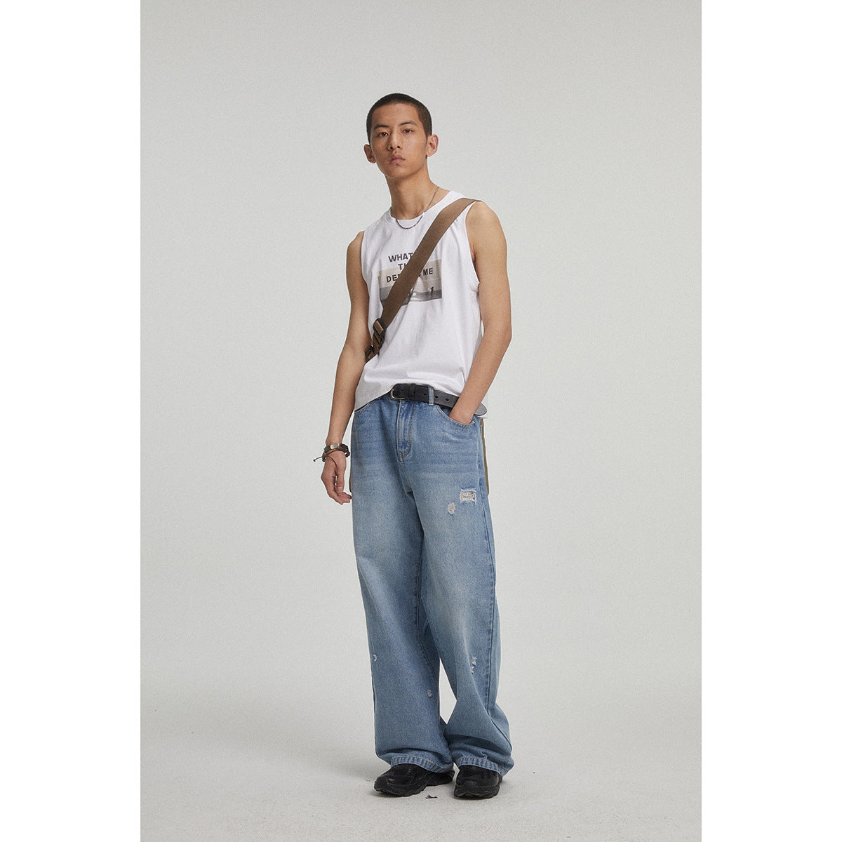 Full-length model wearing a white graphic sleeveless shirt, loose distressed blue jeans and a crossbody strap.
