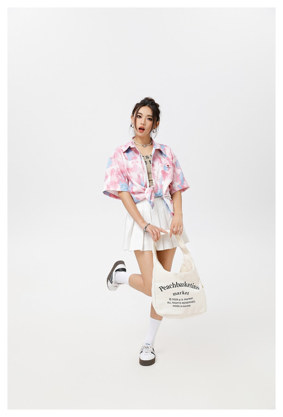 Model wearing a pink and blue abstract-print tied shirt and white pleated skirt, holding a cream printed tote bag.