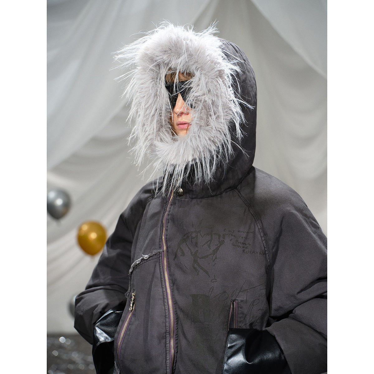 Close-up of a black jacket with a large gray fur-trimmed hood, front zipper, zip pockets and dark printed graphics.
