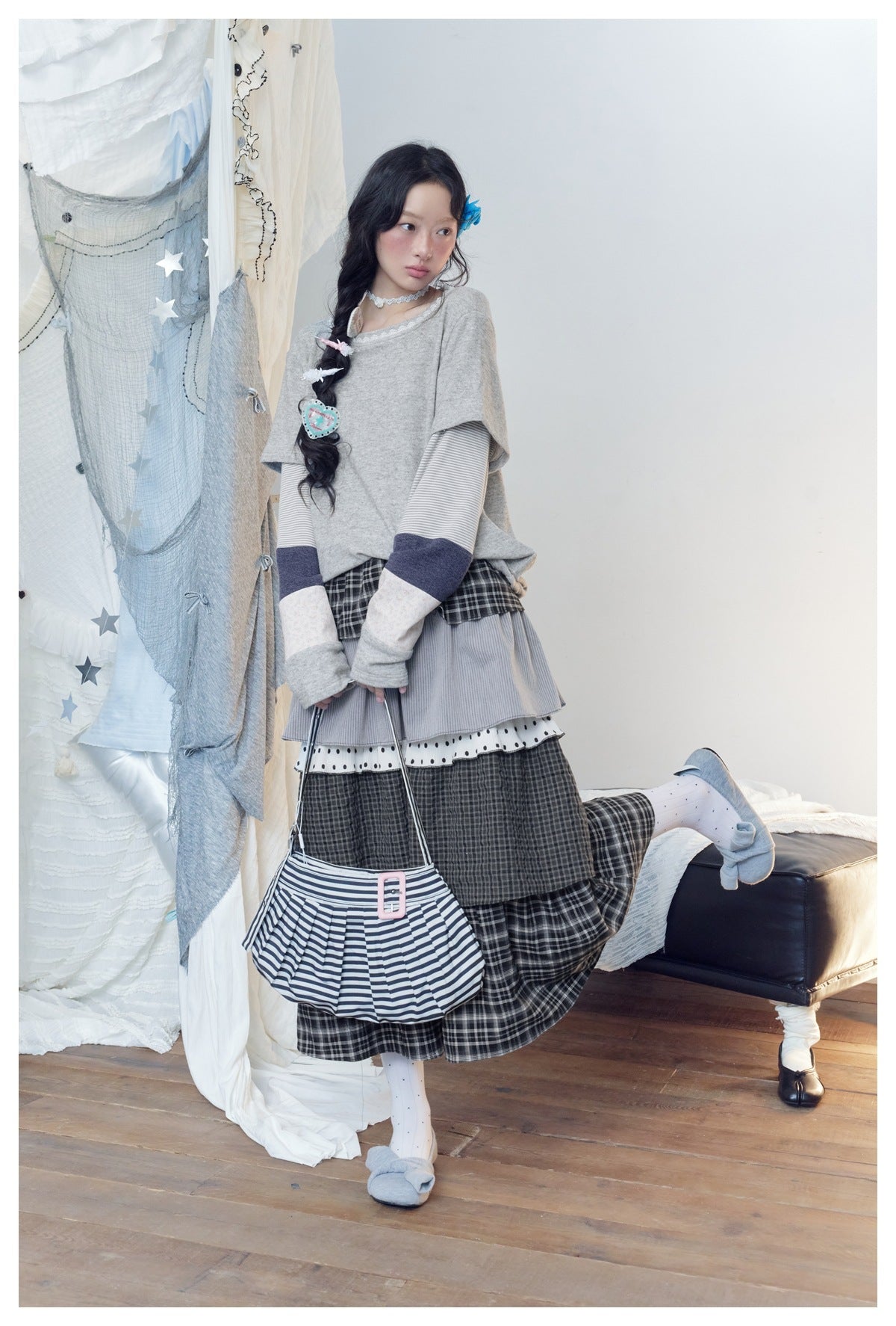 Full-length model wearing a gray layered-sleeve top and tiered plaid skirt with a striped bag.