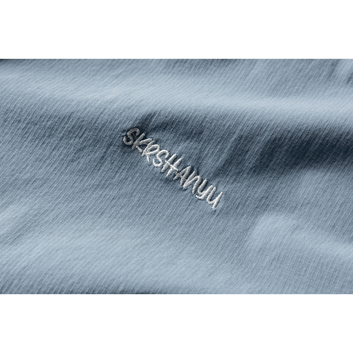 Close-up of white embroidered lettering on textured light blue shirt fabric.