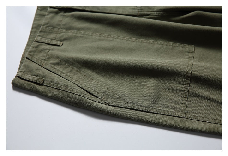 Close-up of olive trousers showing a large rectangular pocket, stitched seams, and small fabric tabs.