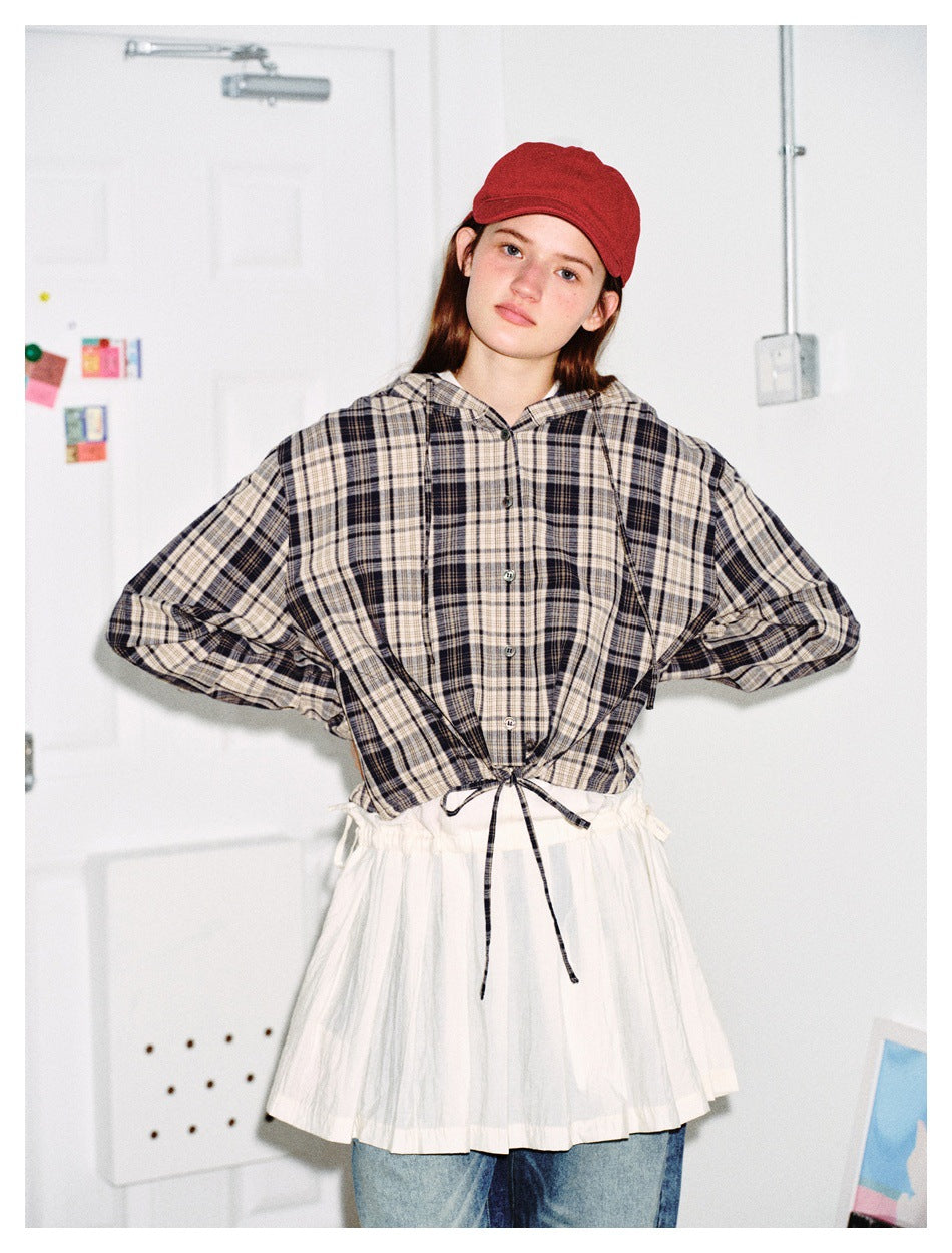 Model wearing a plaid hooded button-up jacket with hem drawstring tied, over a white skirt layer, red cap and jeans