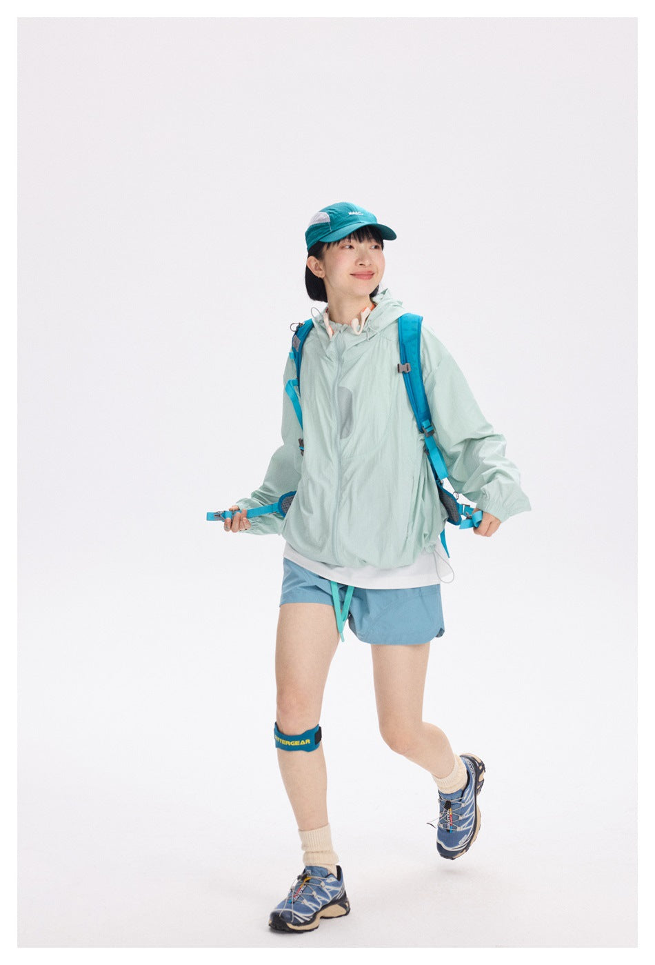 Full-length model wearing a pale mint hooded jacket and blue shorts with a turquoise cap and blue hiking backpack.