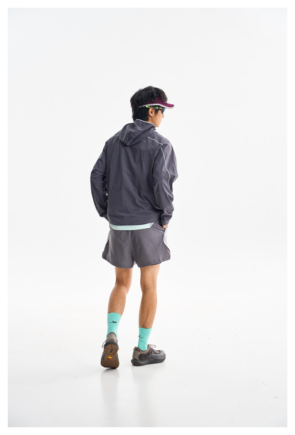 Rear view of a model wearing a charcoal hooded jacket with light piping and gray shorts.