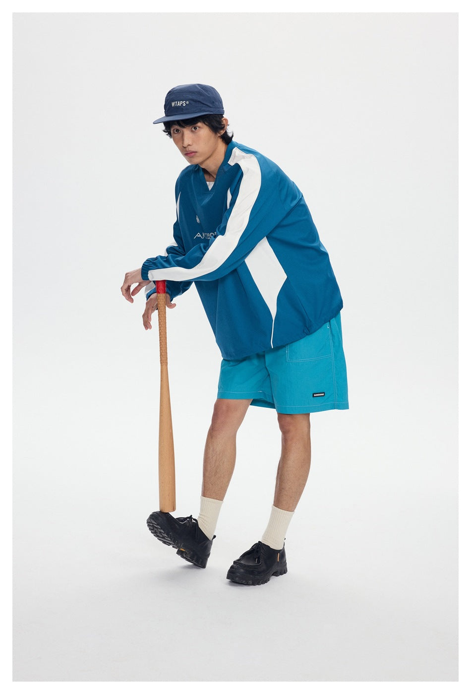 Model wearing a teal-and-white pullover and teal shorts, leaning on a bat against a plain background.