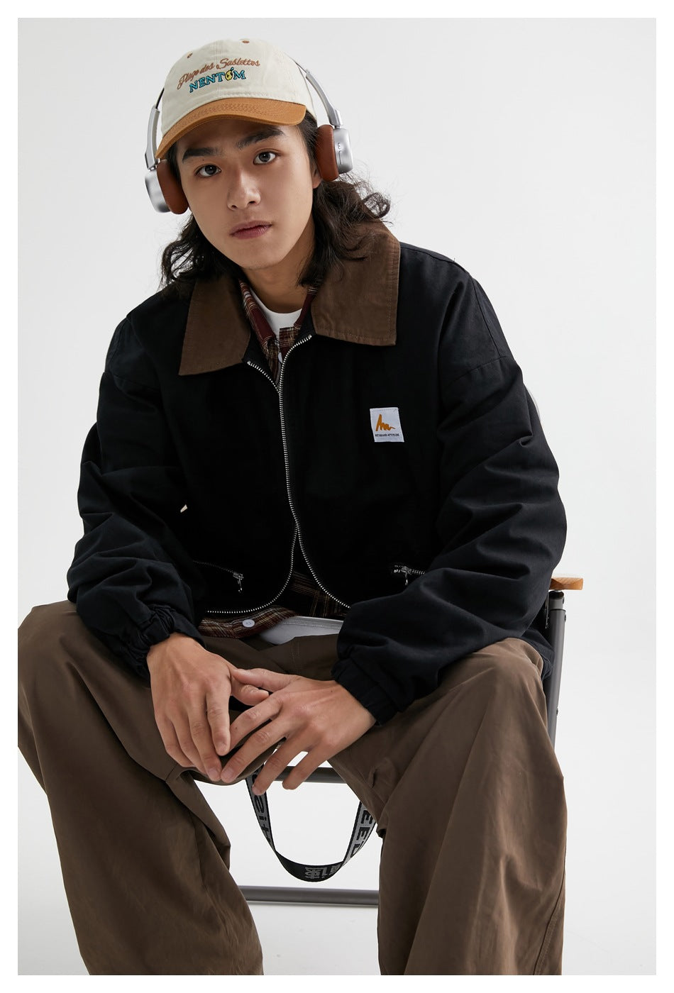 Seated close view of a model wearing a black jacket with a brown collar, silver zipper and zip pockets.