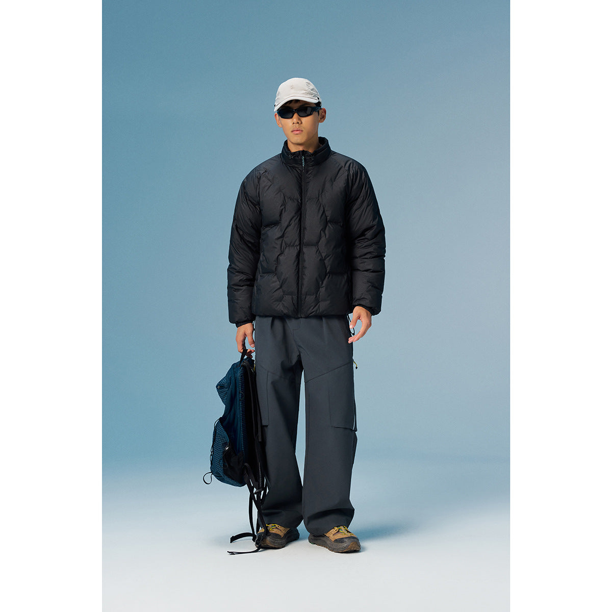 Model wearing a black quilted zip jacket and wide dark cargo pants, with a cap and backpack.