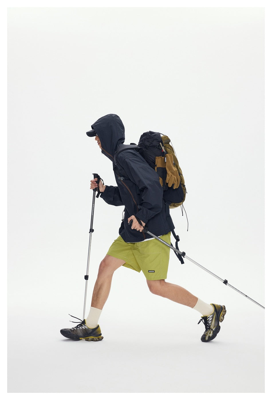 Side view of a model hiking in a dark hooded jacket and lime-green shorts with a backpack and trekking poles.