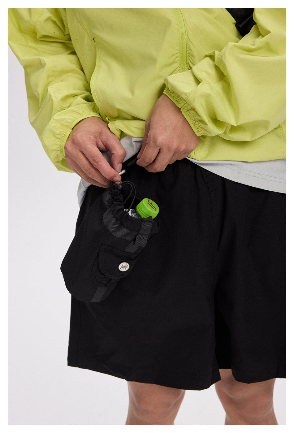 Close-up of a model adjusting the attached black utility pouch on black shorts; the pouch holds a small bottle.