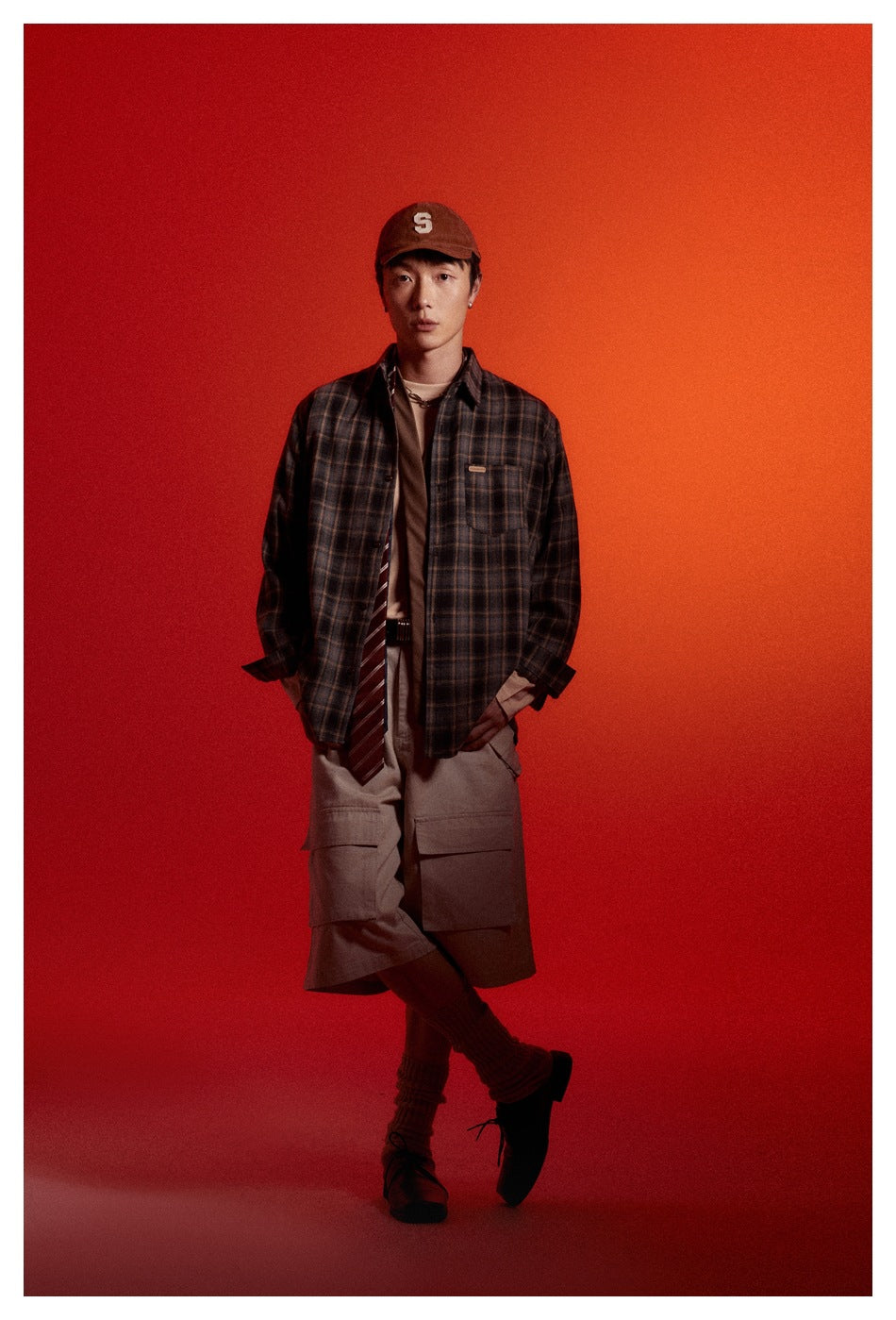 Man wearing an open gray plaid button-up shirt over a tee with cargo shorts and a cap against a red backdrop