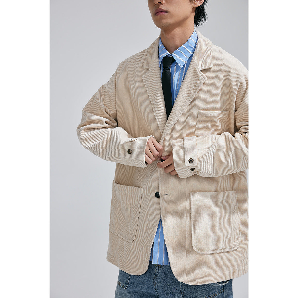 Close view of a model wearing a cream ribbed blazer with patch pockets, buttoned cuffs, a blue striped shirt and black tie.