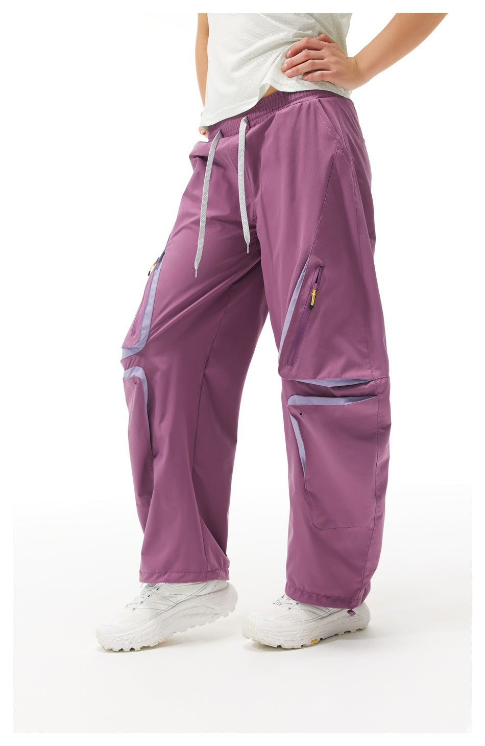 Angled view of a model wearing plum-purple cargo pants with an elastic waist, drawcords, zip pockets and pale trim.
