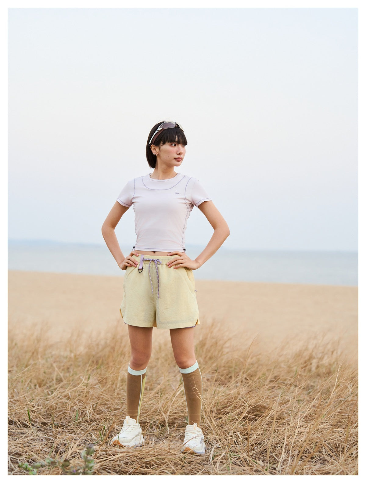 Woman wearing a fitted pale lavender short-sleeve top and pale yellow shorts by the beach.
