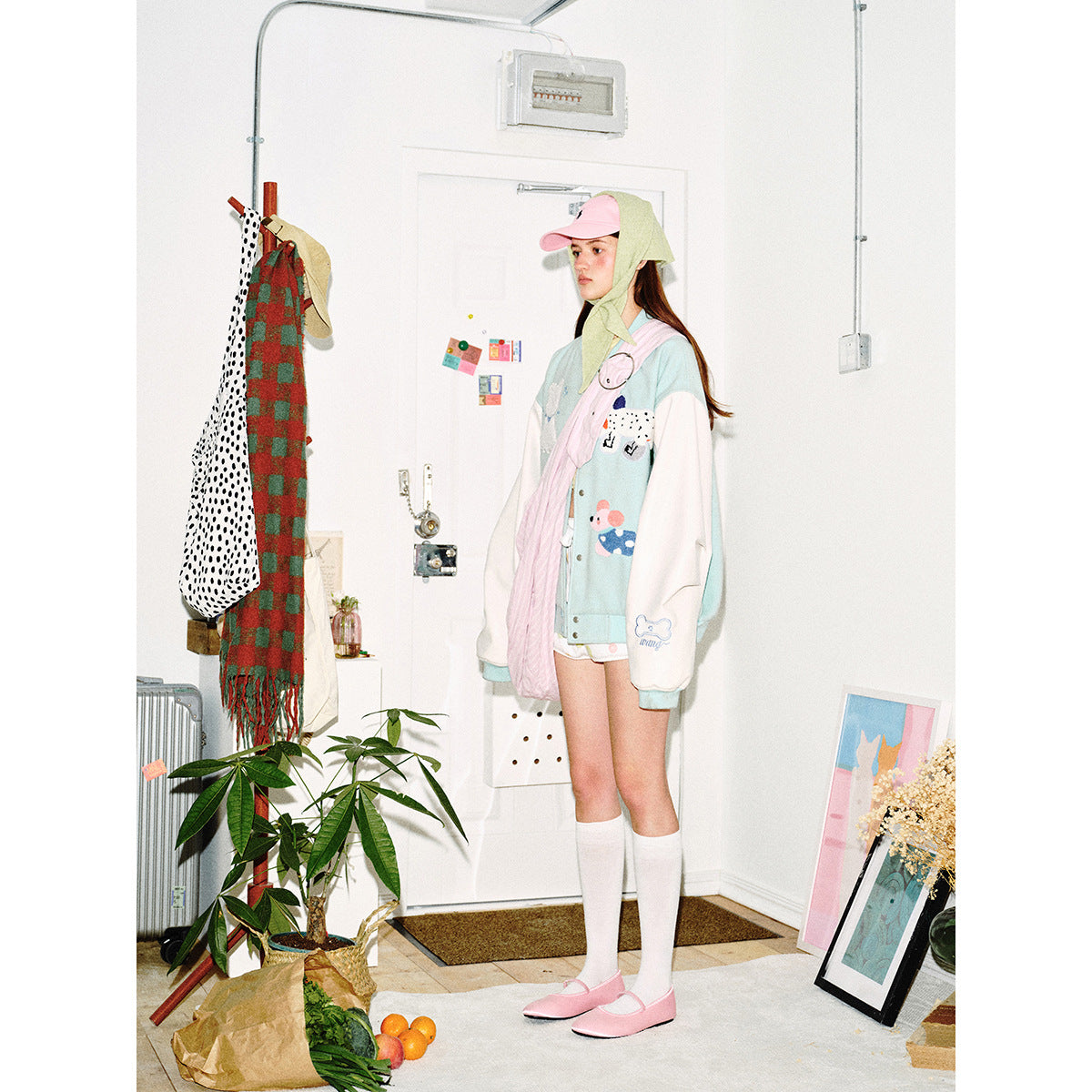 Model in a pale aqua animal-patch varsity jacket with white sleeves, pink shoulder bag, pink cap and white knee socks.