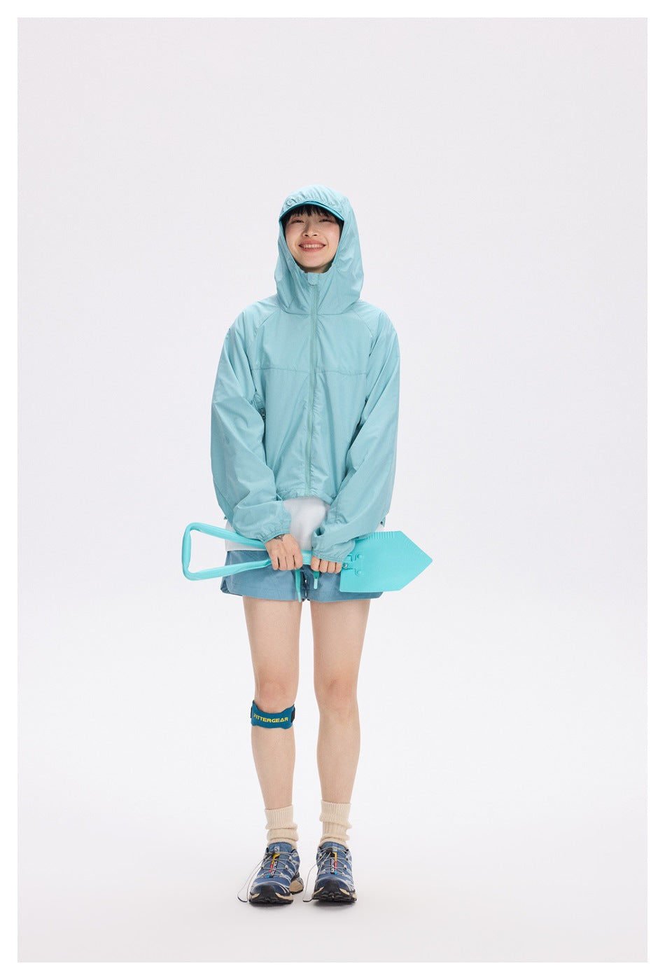 Full-length model wearing a light turquoise hooded jacket and shorts, holding a matching turquoise accessory.