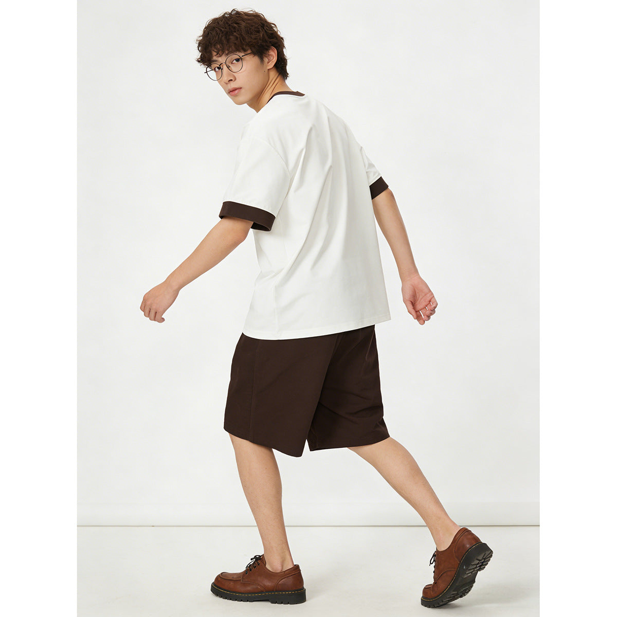 Model showing the back and side of a white short-sleeve T-shirt with dark brown trim, paired with dark shorts.