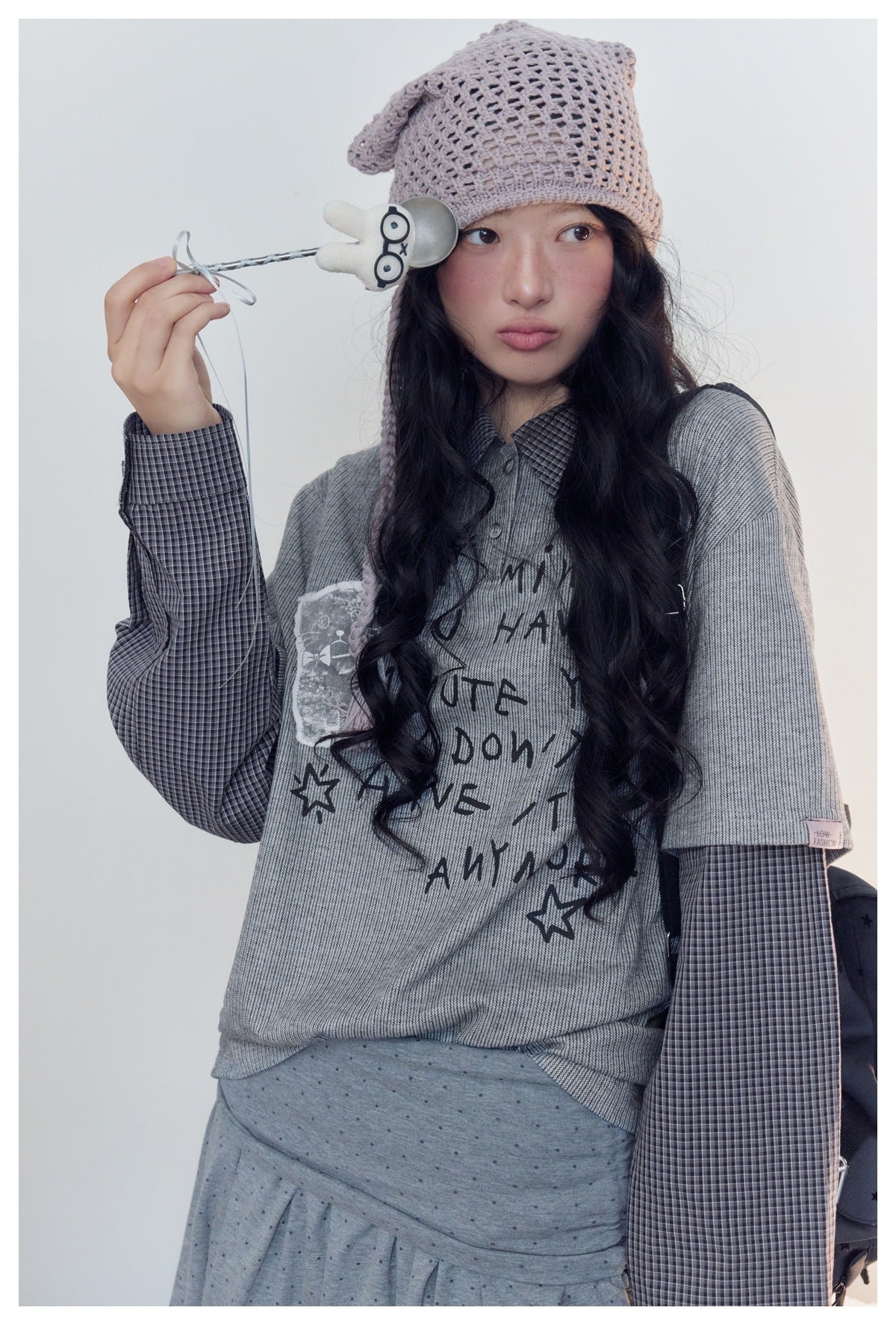 Model wearing a gray graphic top over a dark checked shirt and a gray dotted skirt, with a pale pink crocheted cap.