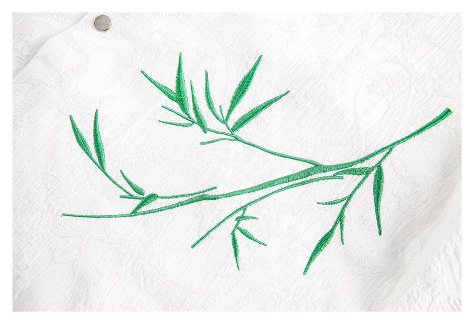 Close-up of a green embroidered leafy branch on white textured fabric, with a metal button near the edge.