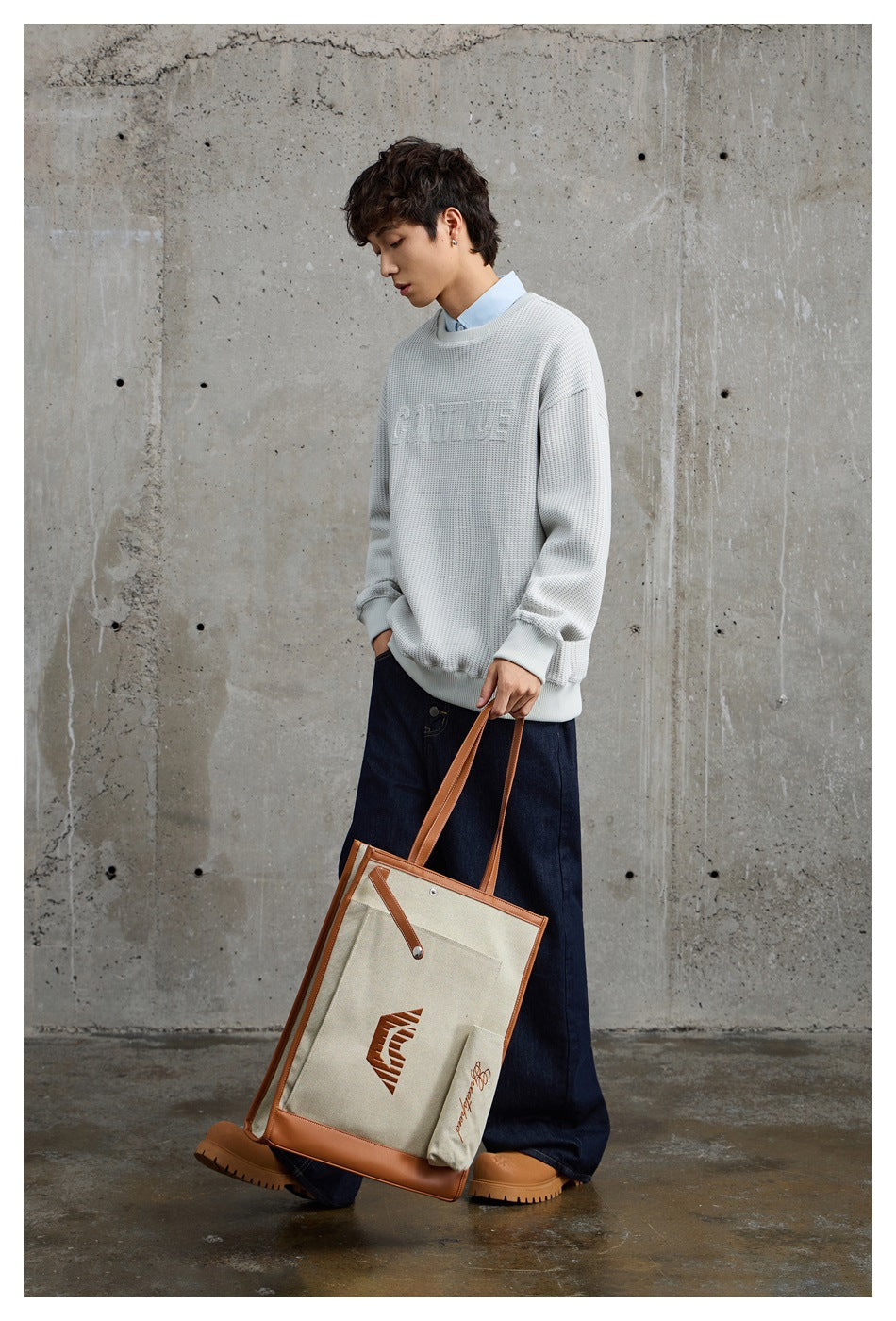 Model wearing a light gray crewneck sweatshirt with embossed lettering and dark wide-leg jeans, carrying a canvas tote