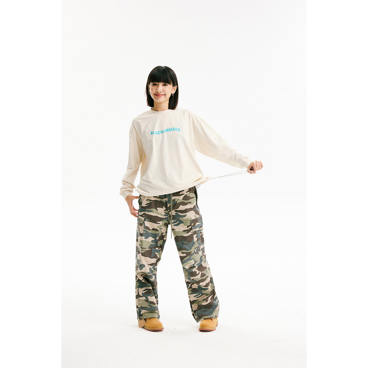 Model wearing a cream long-sleeve shirt with cyan chest lettering and loose camouflage cargo pants.
