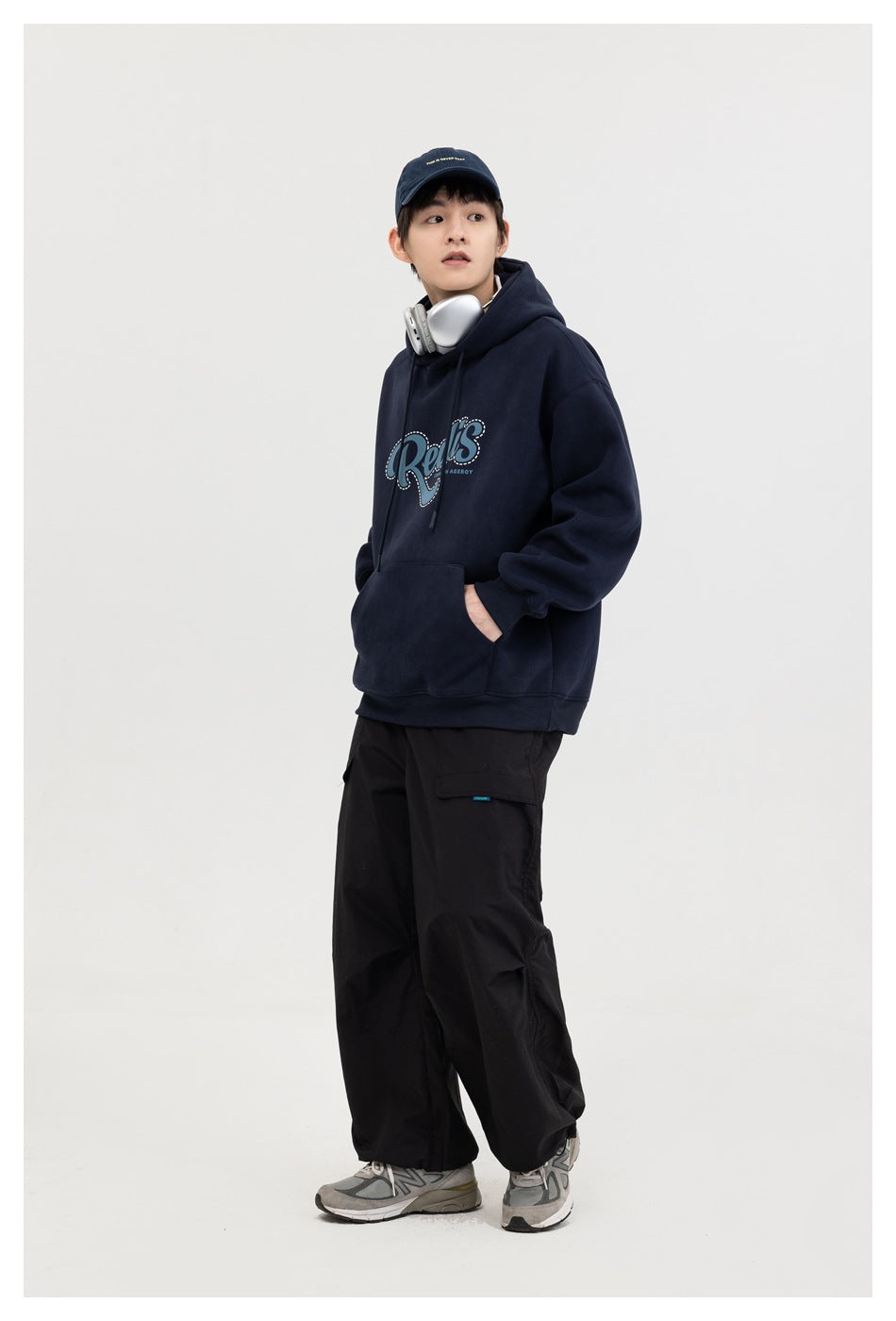 Model wearing a navy hooded sweatshirt with blue script print, hand in pocket, with black cargo pants and sneakers