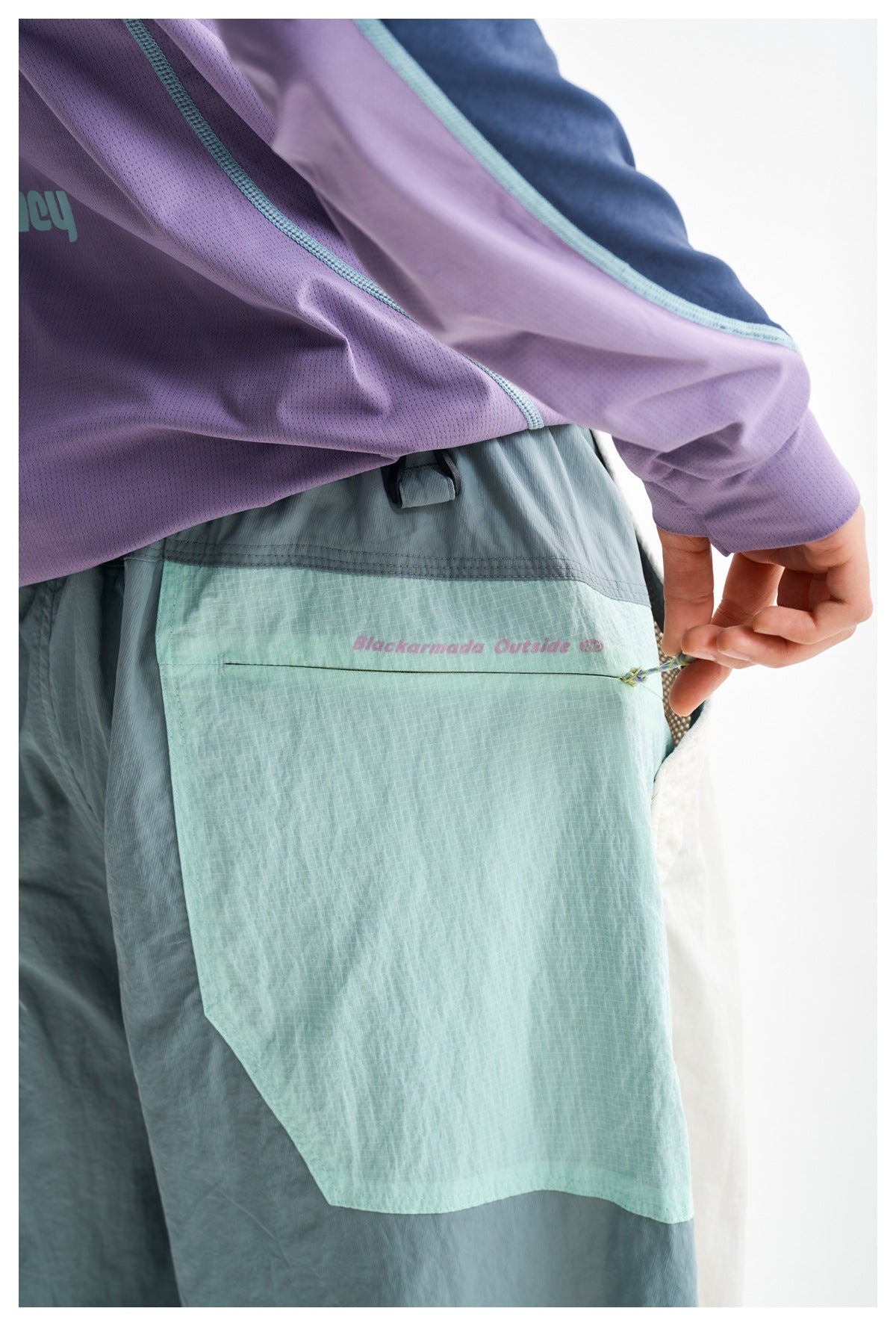 Close-up of mint-green trousers with a large pocket as a hand pulls its zipper cord; a lavender sleeve is visible above.