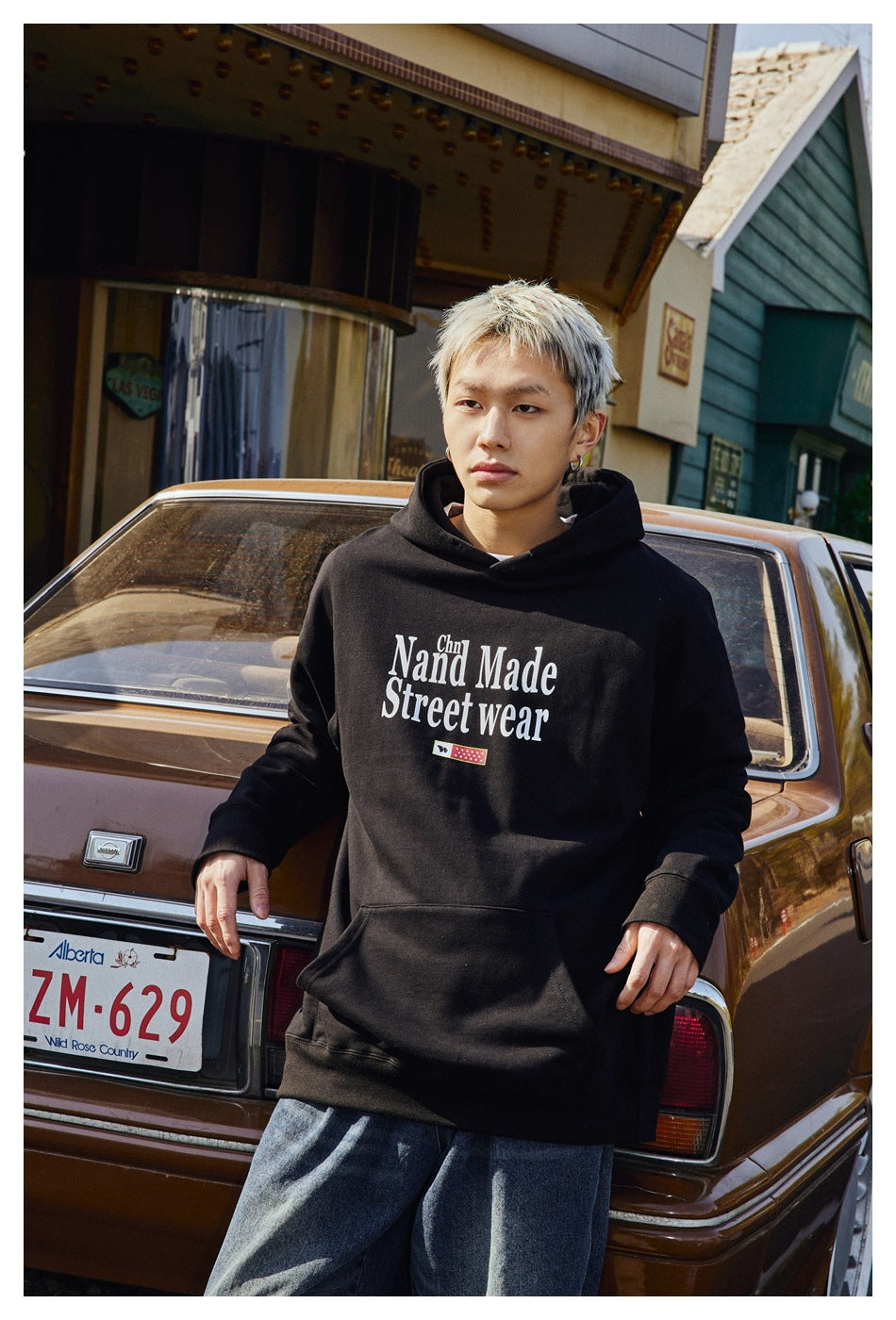 A man wears a black hoodie with white “Hand Made Street wear” lettering and loose blue jeans beside a vintage car.