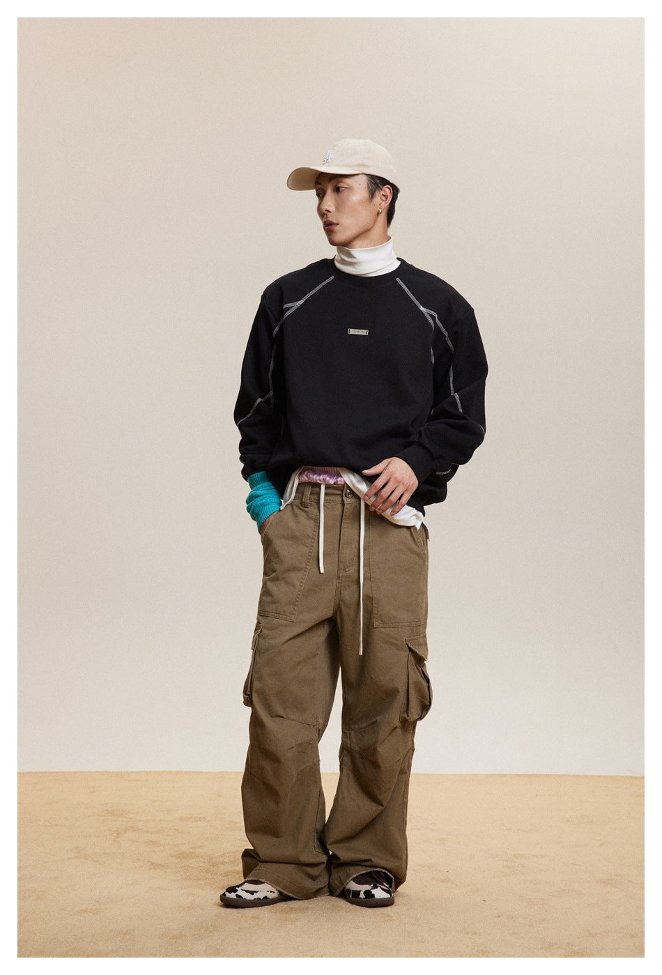 Model wearing a black topstitched crew-neck sweatshirt over a white turtleneck with khaki cargo pants and a cap