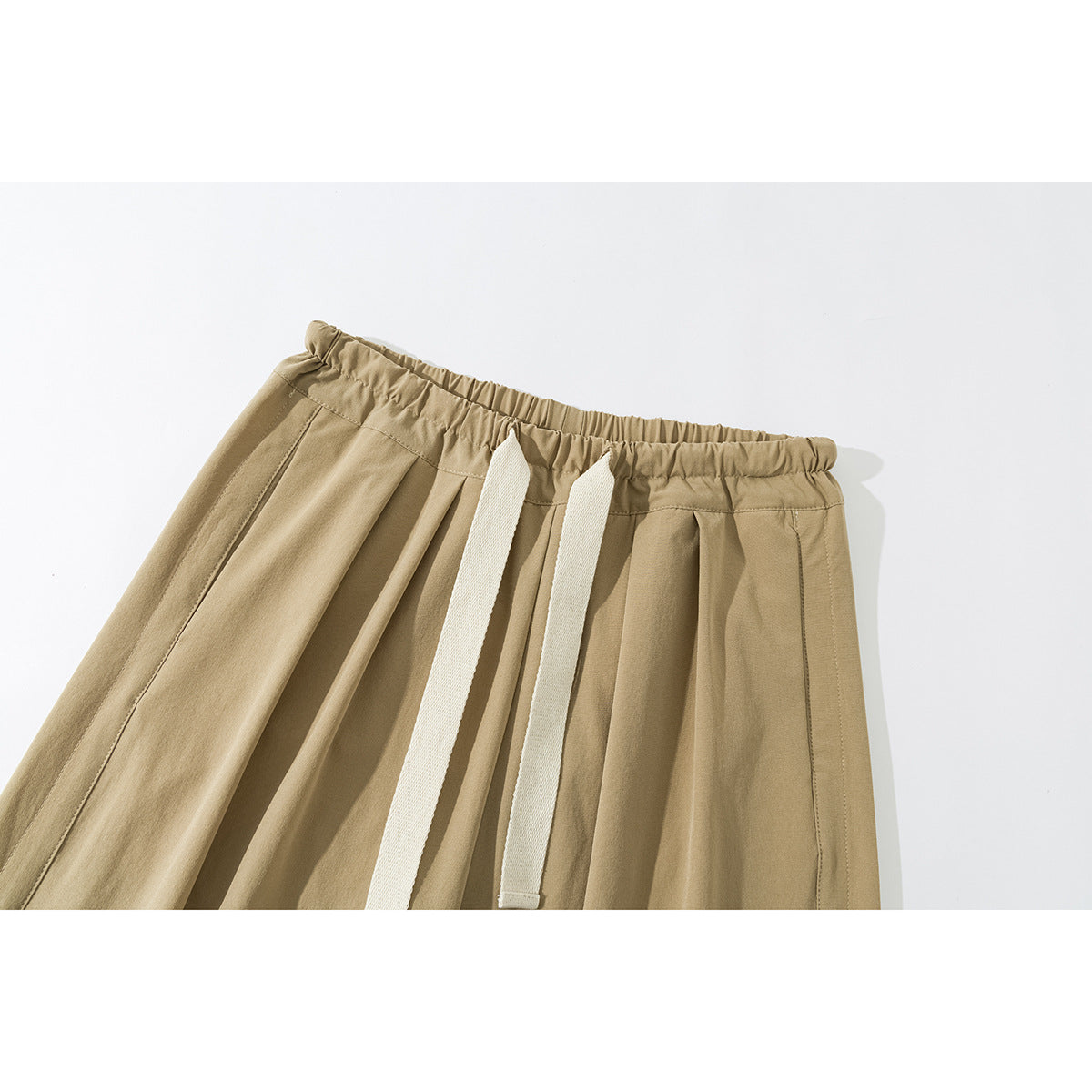 Close-up of tan trousers showing the gathered elastic waistband, front pleats, and two long cream drawstrings.
