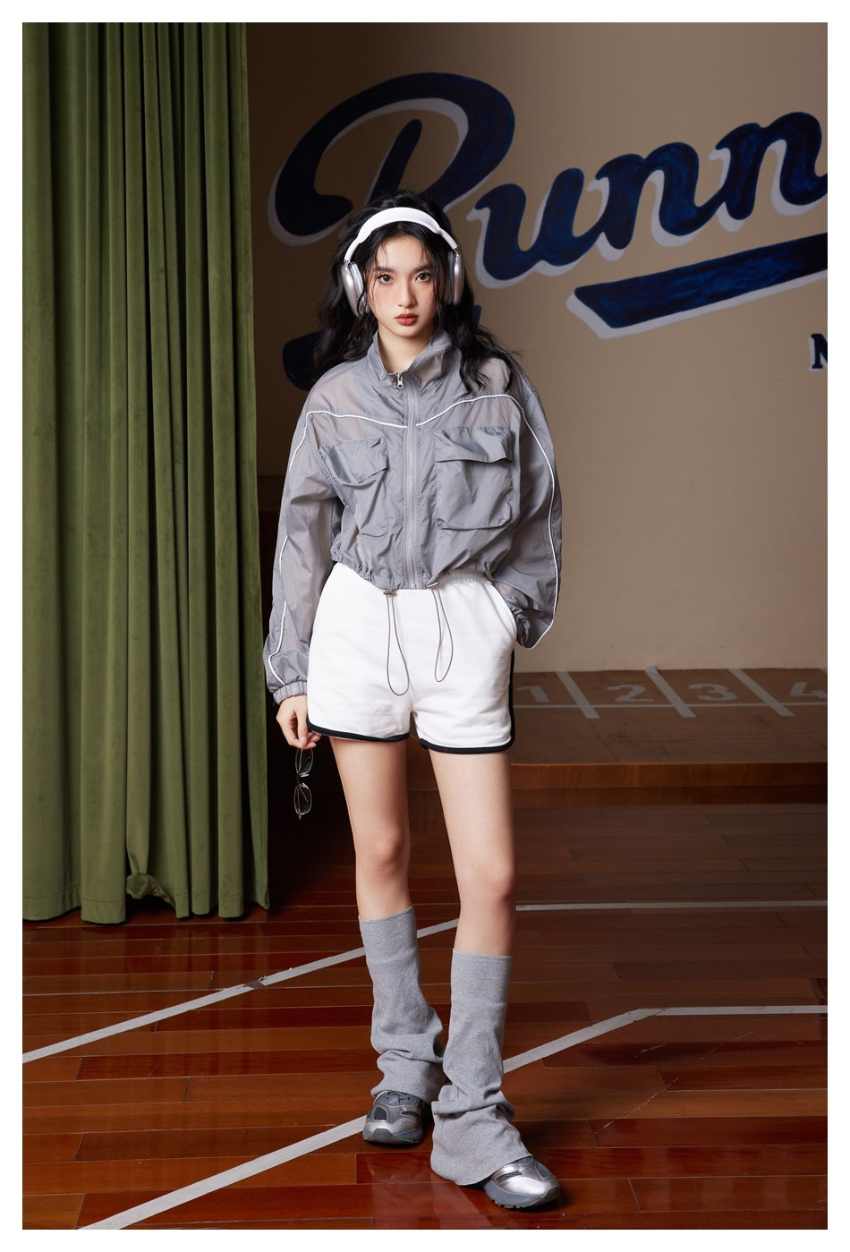 Model wearing cropped gray zip jacket with white piping over white shorts, gray leg warmers and headphones