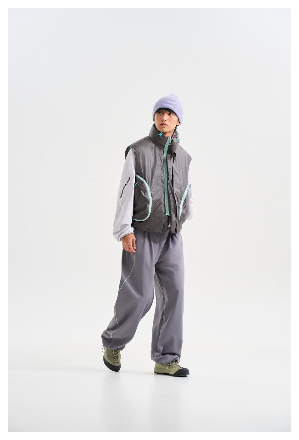 Model wearing a glossy charcoal sleeveless vest with aqua trim, white sleeves, loose gray trousers and a lavender beanie.