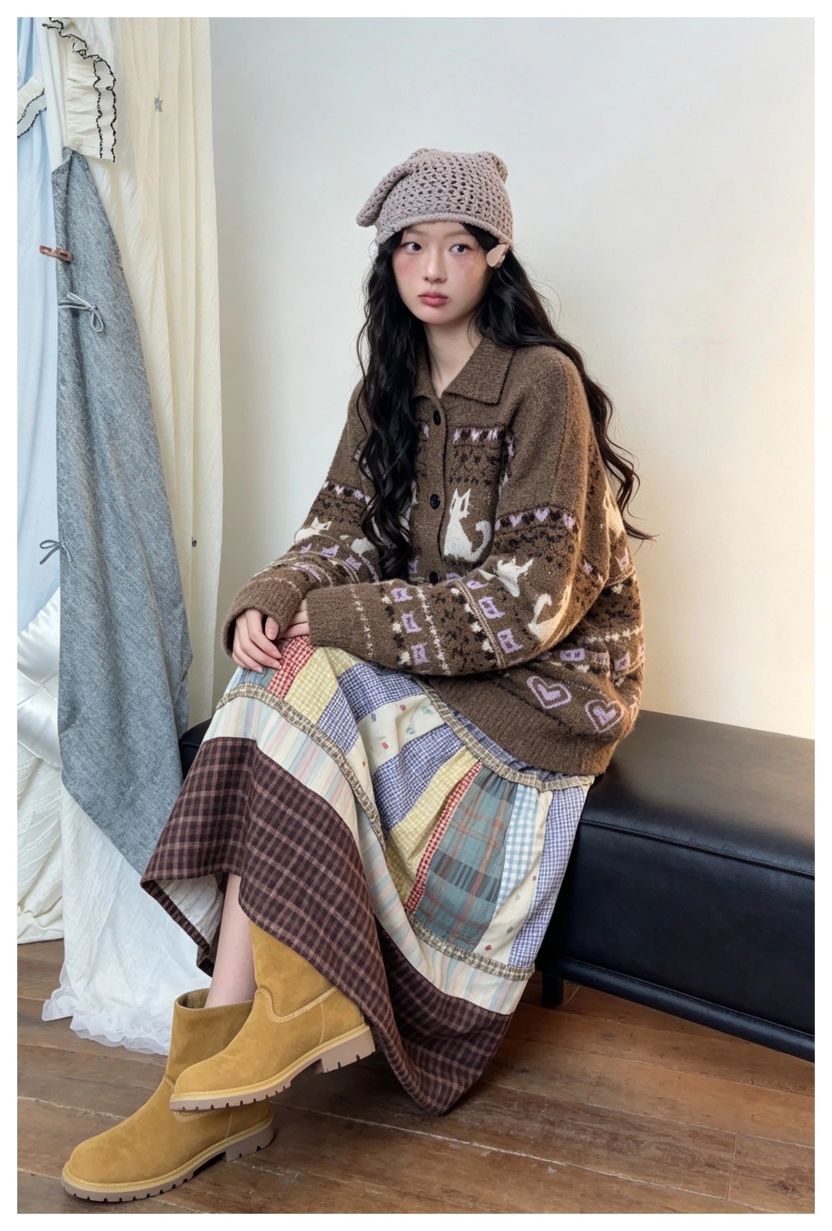 Woman seated wearing a brown cat and heart jacquard cardigan with a patchwork plaid skirt, beanie and yellow boots