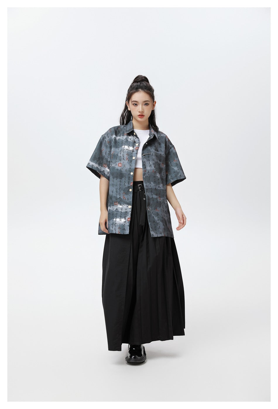 Model wearing the black-gray calligraphy-print short-sleeve shirt open over a white crop top with a long black pleated skirt