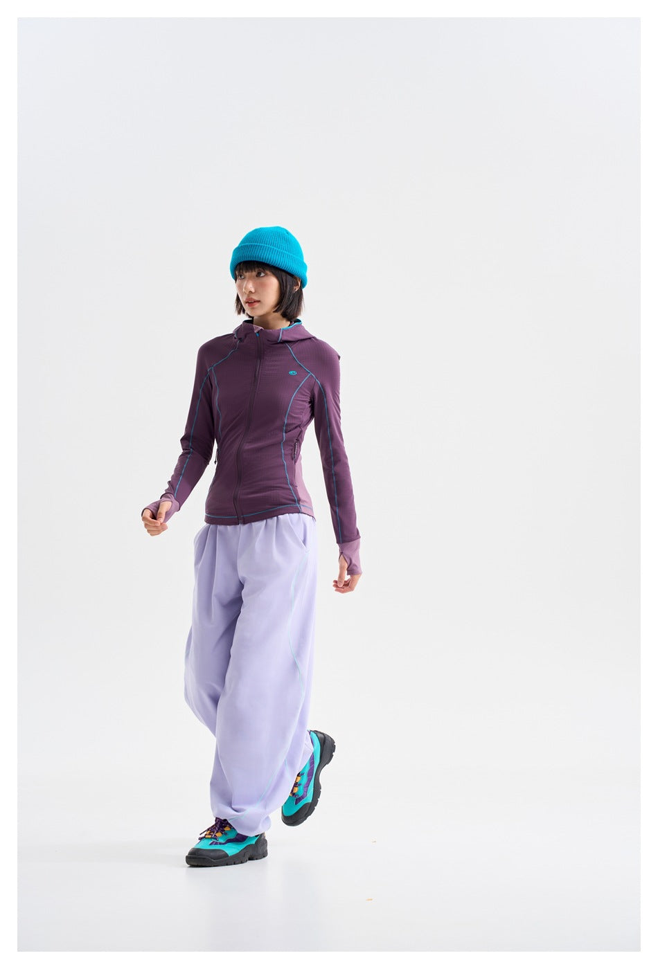 Model walking in a purple zip-up hooded jacket with turquoise piping and loose lavender pants, wearing a bright blue beanie.