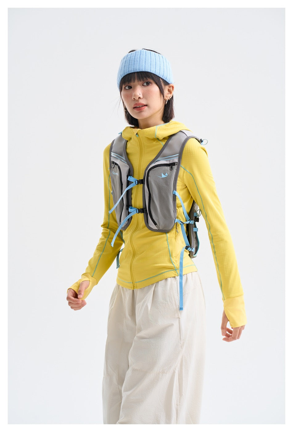 Model wearing a yellow hooded zip jacket with turquoise seams, a gray hiking vest, and loose white pants.
