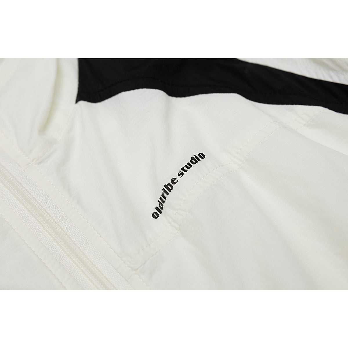 Close-up of an ivory jacket showing a small black “oldtribe studio” chest print and a light-colored zipper.