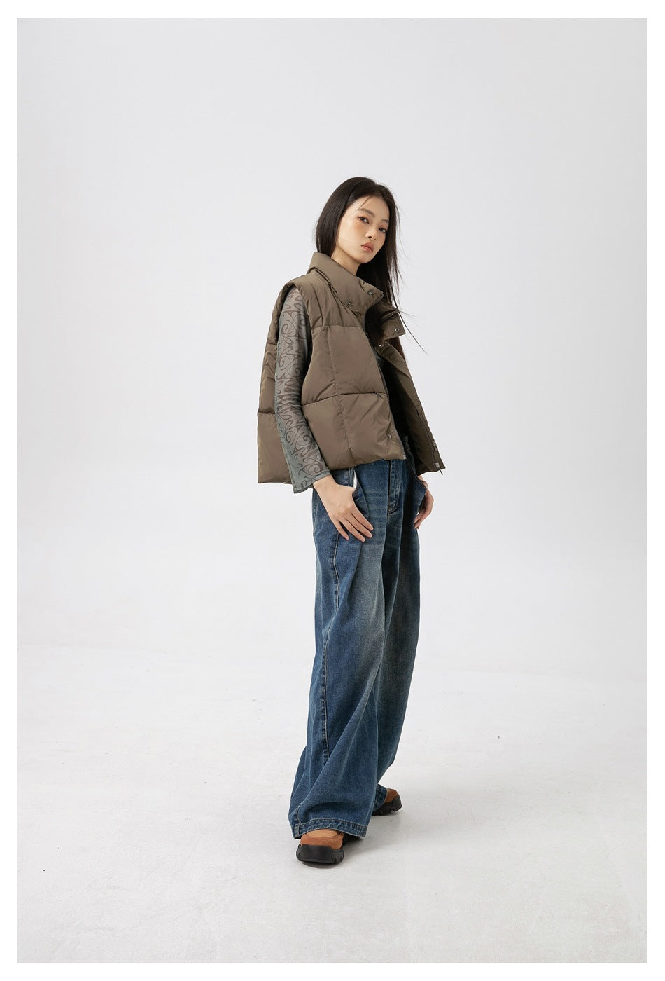 Full-length model wearing a taupe quilted puffer vest over a gray patterned long-sleeve top and wide-leg jeans.