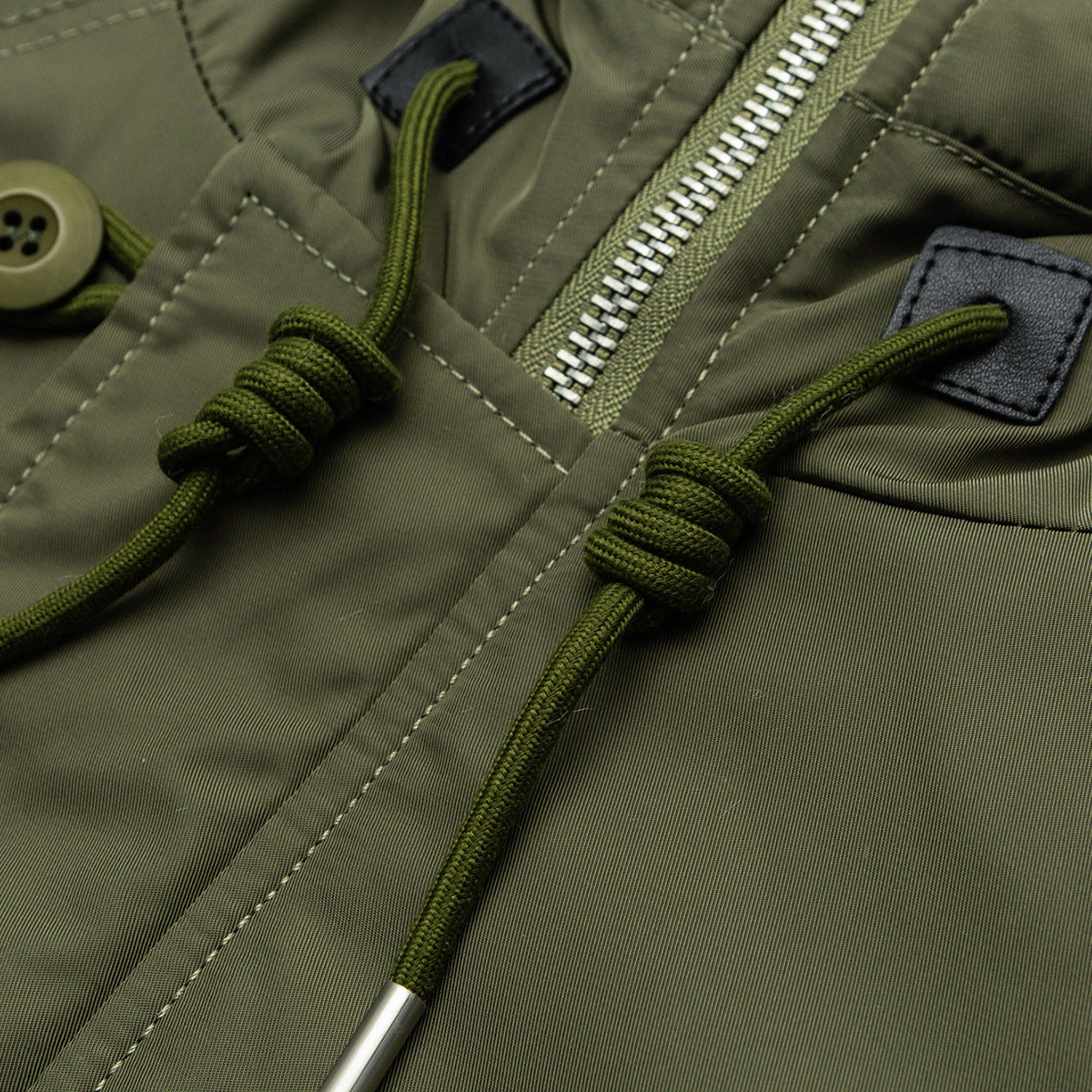 Close-up of an olive parka showing a silver zipper, braided drawcords, stitching, and a button.
