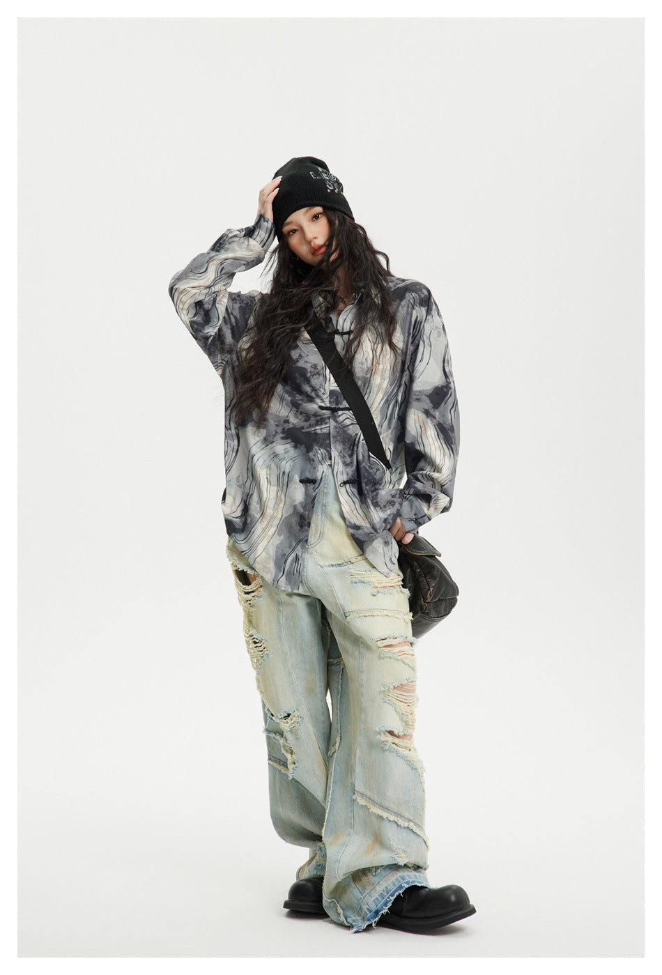 A model poses in a loose gray abstract-pattern shirt and heavily distressed light jeans with a black beanie.