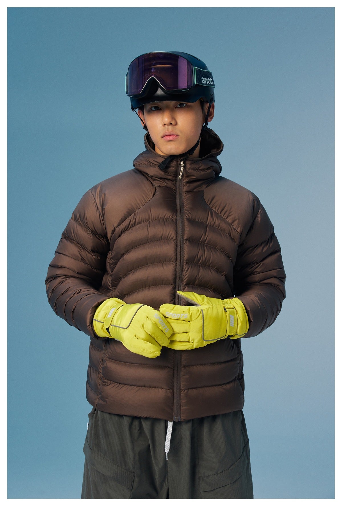 Model wearing a dark brown hooded puffer jacket and neon yellow gloves, with ski goggles and helmet.